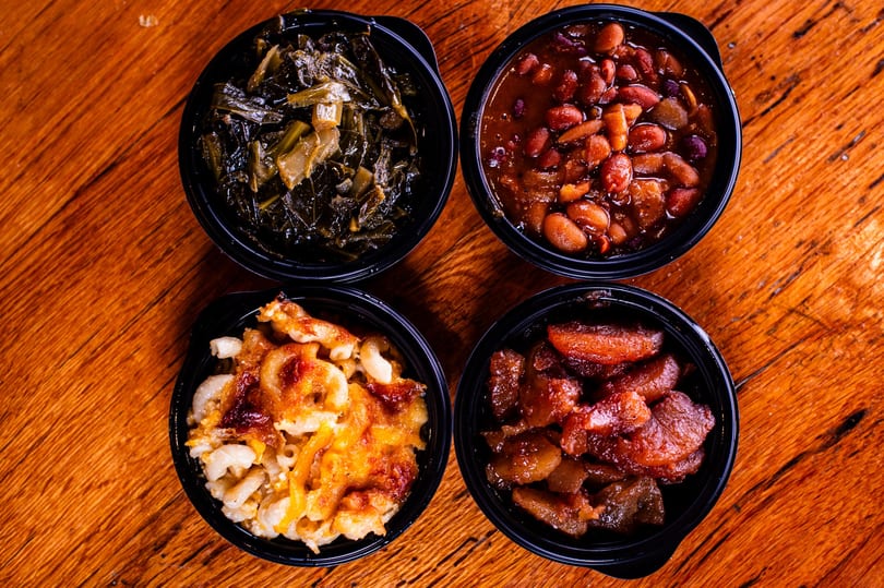 Four black bowls filled with collard greens, baked beans, macaroni and cheese, and candied yams.