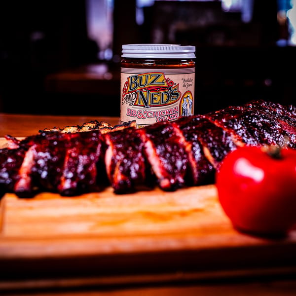 Sliced BBQ ribs, Buz and Ned's glaze, and a tomato on a wooden cutting board.