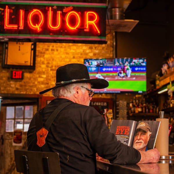 Man in a cowboy hat reading a magazine at a bar with a neon "LIQUOR" sign.