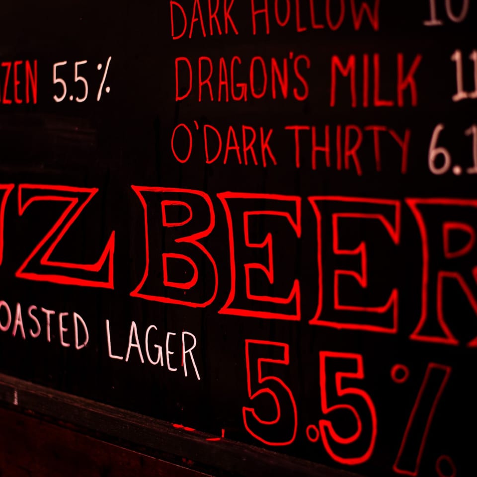 Chalkboard menu with red and white text listing beer names and alcohol percentages.