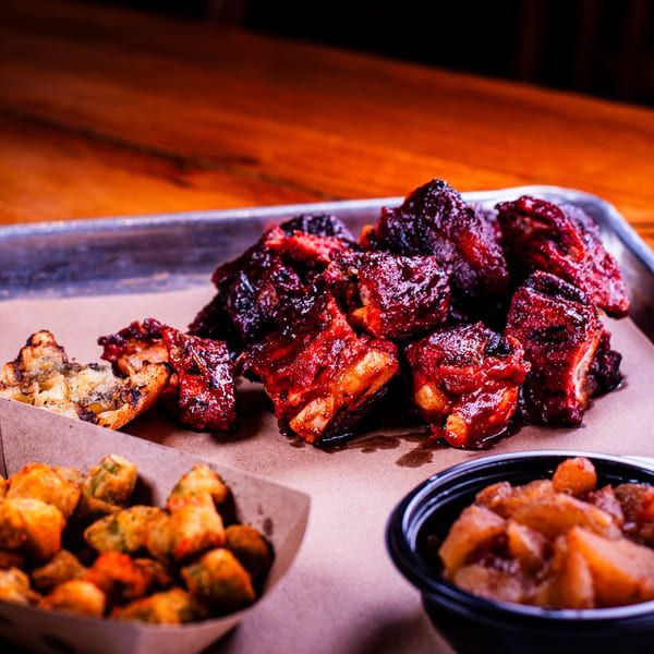 A metal tray with saucy BBQ ribs, fried okra, and baked apples on a wooden table.