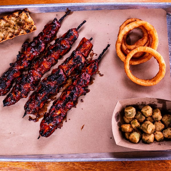 Four glazed BBQ skewers, grilled bread, crispy onion rings, and fried okra on a metal tray.