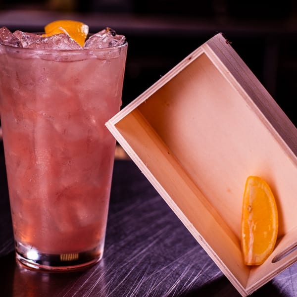 Pink cocktail with ice and orange peel, next to a wooden box with an orange slice.