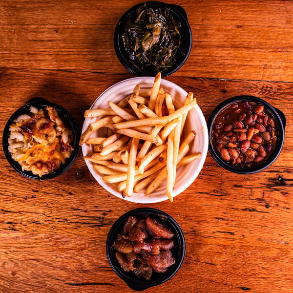 Assortment of sides including fries, mac and cheese, collard greens, baked beans, and candied yams on a wooden table.
