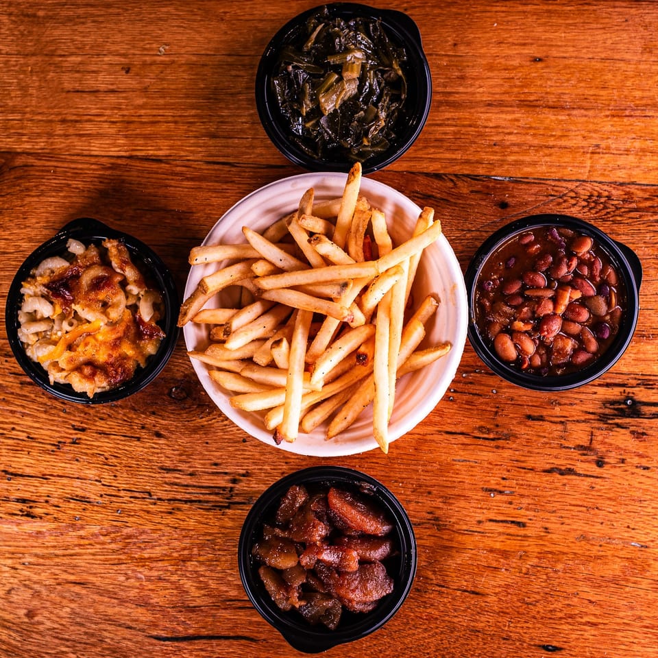 Assortment of sides including fries, mac and cheese, collard greens, baked beans, and candied yams on a wooden table.